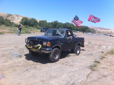 1990 Ford Bronco - A dark-colored Ford Bronco is parked on a dirt path, with two flags waving in the wind behind it.