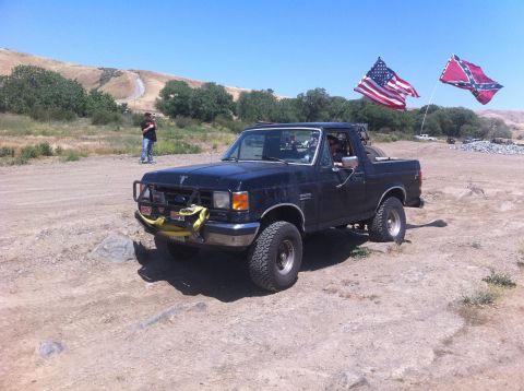 1990 Ford Bronco - A black Ford Bronco is parked off-road with two flags flying from poles on its rear.