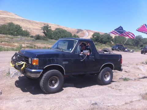 1990 Ford Bronco - A black Ford Bronco is parked off-road with American and Confederate flags flying.