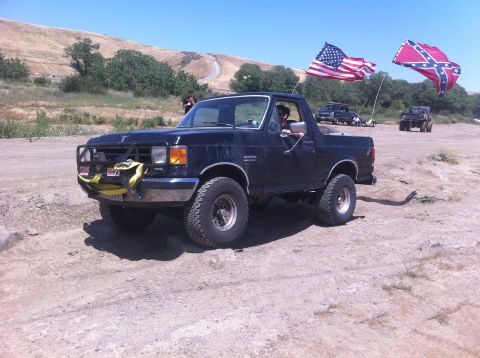 1990 Ford Bronco - A dark-colored Ford Bronco is parked on a dusty trail with two flags flying in the background.