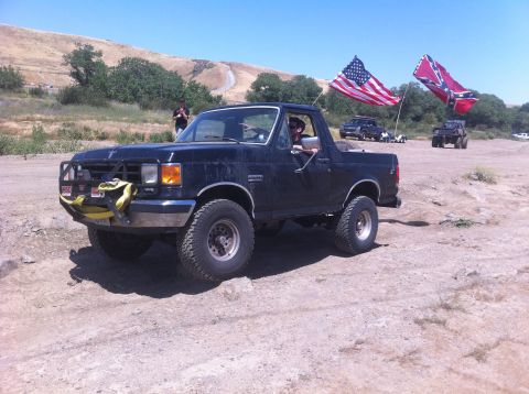 1990 Ford Bronco - A black Ford Bronco is parked off-road, displaying American and Confederate flags.