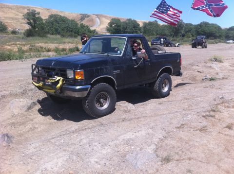 1990 Ford Bronco - A black Ford Bronco is shown off-roading with two flags flying in the background.