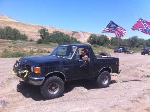 1990 Ford Bronco - A black Ford Bronco is parked on a dirt road with American and Confederate flags flying.