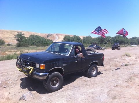 1990 Ford Bronco - A black 1990 Ford Bronco drives off-road with two flags displayed, joined by other vehicles in the background.