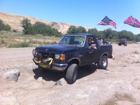 1990 Ford Bronco - A black Ford Bronco is shown off-roading with American and Confederate flags flying.