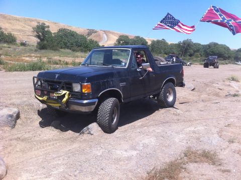 1990 Ford Bronco - 1990 Ford Bronco navigates rough terrain with American and Confederate flags flying.