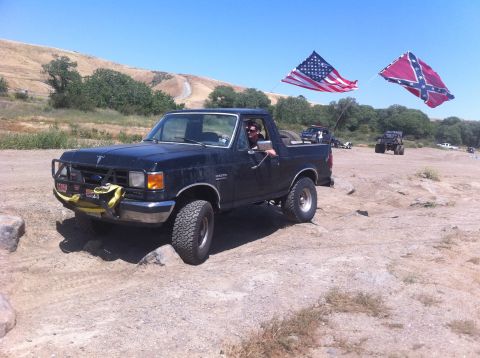 1990 Ford Bronco - A black Ford Bronco is shown off-roading with two flags flying from its rear.