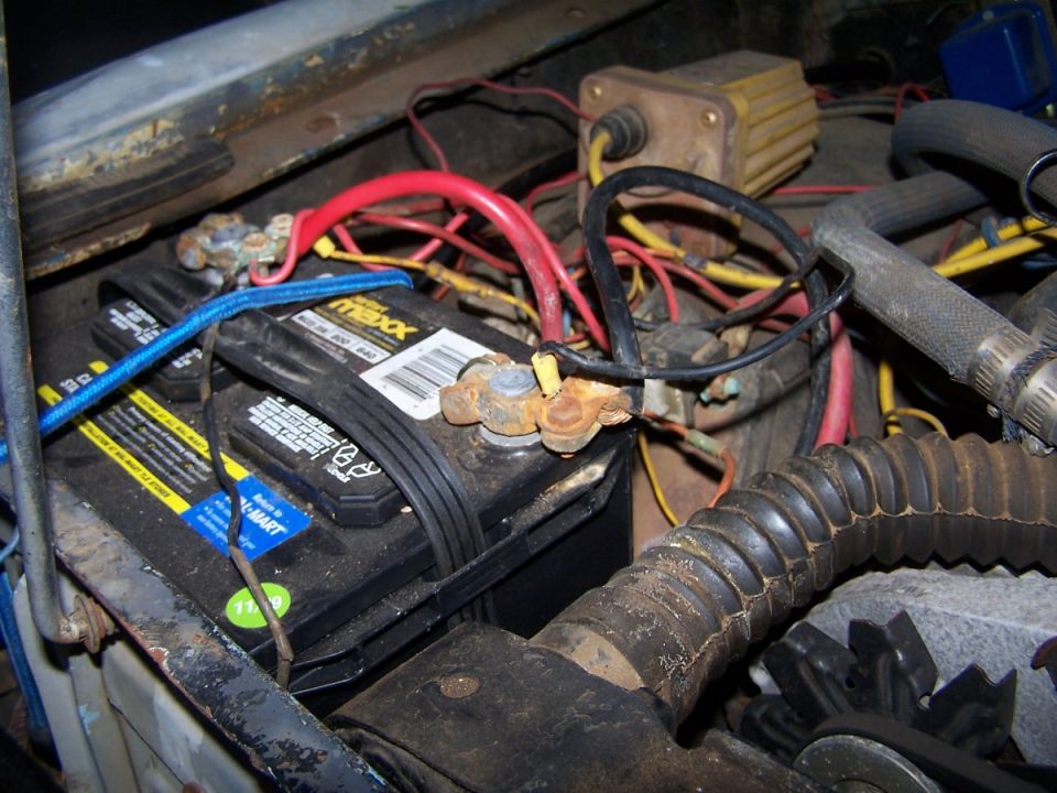 Engine bay showing a black car battery with red and blue cables, rusty terminals, and various wires