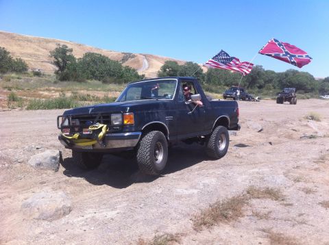 1990 Ford Bronco - A black Ford Bronco drives off-road, flying the American and Confederate flags.
