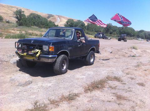 1990 Ford Bronco - A black Ford Bronco drives off-road with two flags flying from its rear.