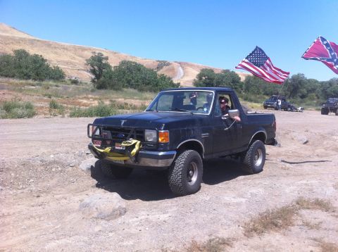 1990 Ford Bronco - Black 1990 Ford Bronco navigates rough terrain with American and Confederate flags flying.