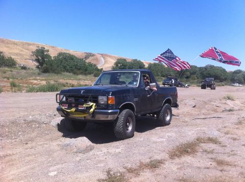1990 Ford Bronco - A black 1990 Ford Bronco is seen off-roading with two flags flying from its rear.