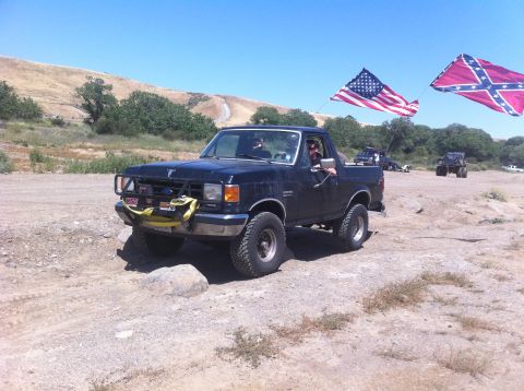 1990 Ford Bronco - A black Ford Bronco is shown off-road with two flags flying from poles attached to the vehicle.
