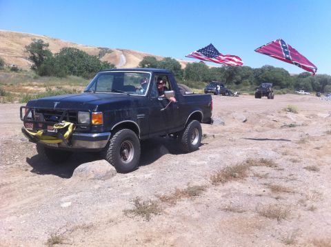 1990 Ford Bronco - A black 1990 Ford Bronco navigates rough terrain, with American and Confederate flags visible in the background.