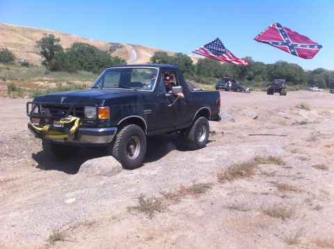 1990 Ford Bronco - Ford Bronco navigates rough terrain under a clear blue sky, with American and Confederate flags in the background.