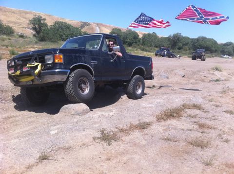 1990 Ford Bronco - A 1990 Ford Bronco navigates rough terrain, with other vehicles and flags visible in the background.