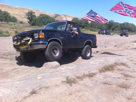 1990 Ford Bronco - A dark blue Ford Bronco navigates a rocky off-road trail, with American and Confederate flags visible in the background.