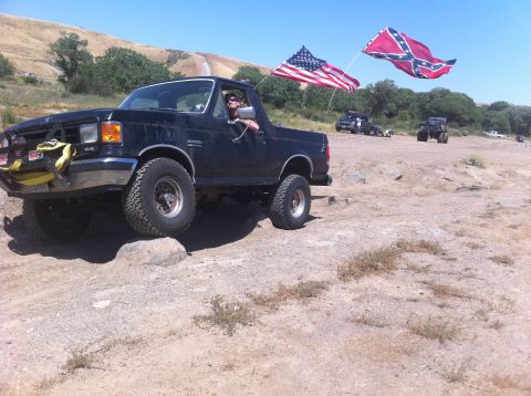 1990 Ford Bronco - A black Ford Bronco navigates rough terrain, flying two flags from its rear.