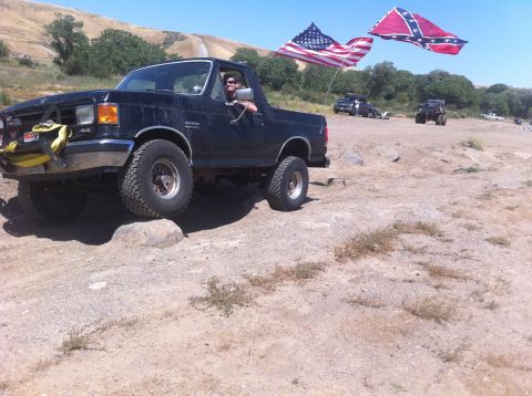 1990 Ford Bronco - A black Ford Bronco navigates rough terrain, flying two flags from its rear.