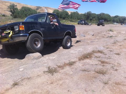 1990 Ford Bronco - This 1990 Ford Bronco navigates a rocky trail under a clear blue sky, with multiple flags visible in the background.