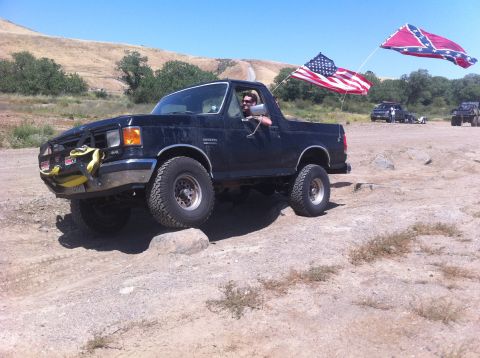 1990 Ford Bronco - Driver waves from a black 1990 Ford Bronco as it navigates an off-road trail with flags flying.