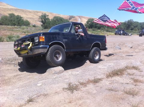 1990 Ford Bronco - A black 1990 Ford Bronco navigates rocky terrain under a clear blue sky, with flags flying in the background.