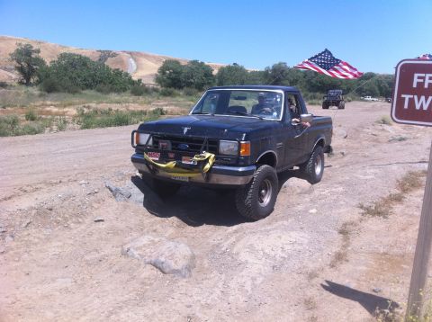 1990 Ford Bronco - A black Ford F-150 pickup truck is driving on a dirt road with an American flag flying from its antenna.