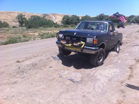 1990 Ford Bronco - A black Ford F-Series truck drives off-road with an American flag flying from its antenna.