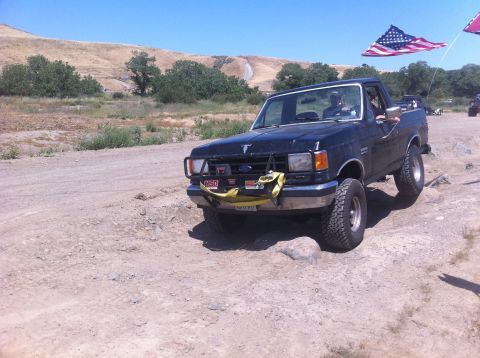 1990 Ford Bronco - A black Ford F-Series truck drives over rocks under a clear blue sky, with American flags flying.
