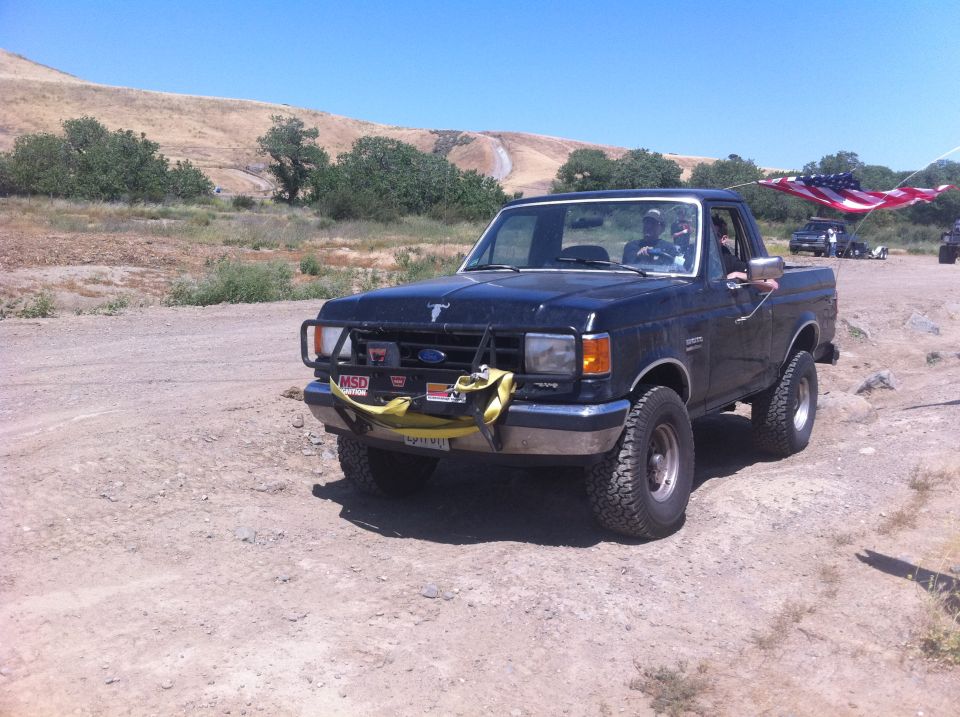 Black Ford F-150 pickup truck with an American flag flying from the antenna, driving on a dirt road