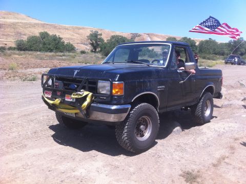 1990 Ford Bronco - A black Ford Bronco drives off-road with an American flag waving in the background.