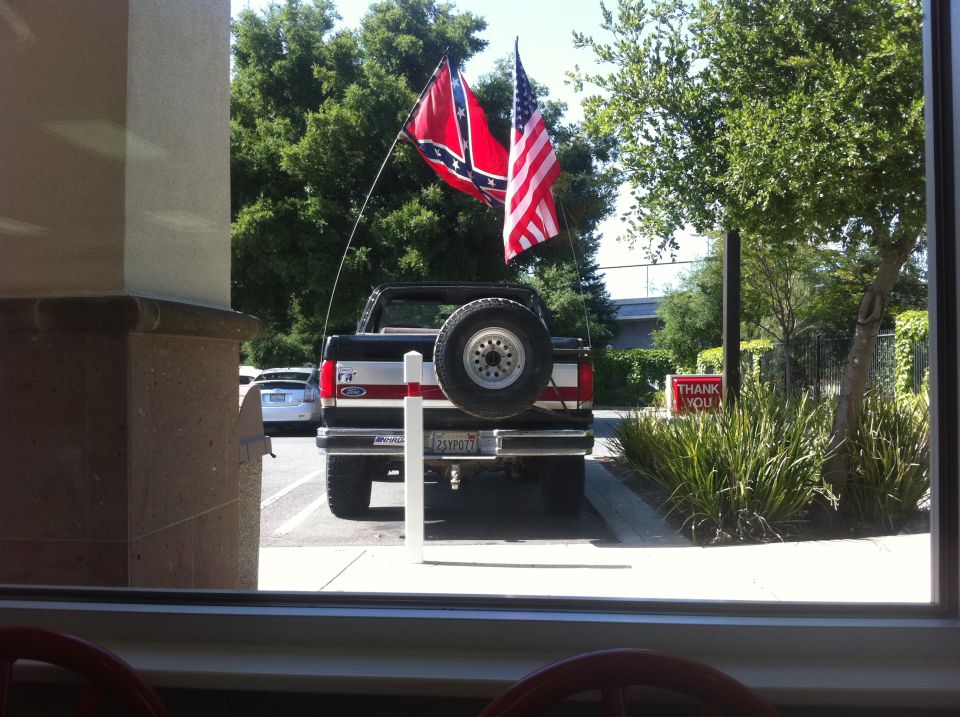 Rear view of a black Ford Bronco with a spare tire mounted on the back, flying a Confederate flag and an American flag