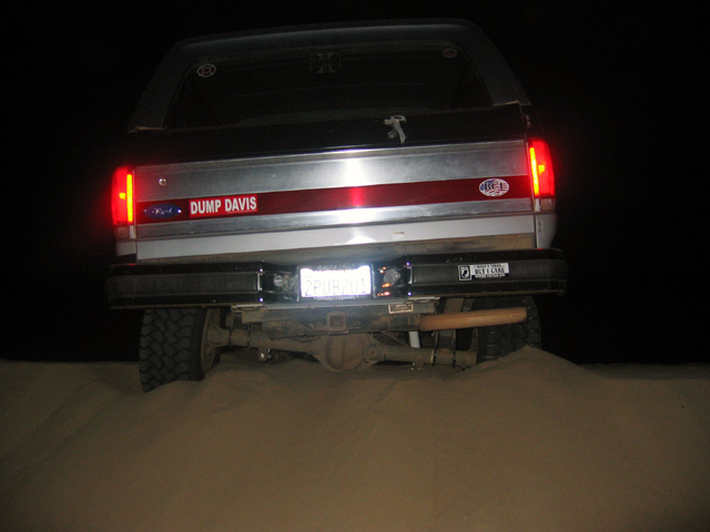 Rear view of a silver Ford Bronco on a sand dune at night with taillights lit