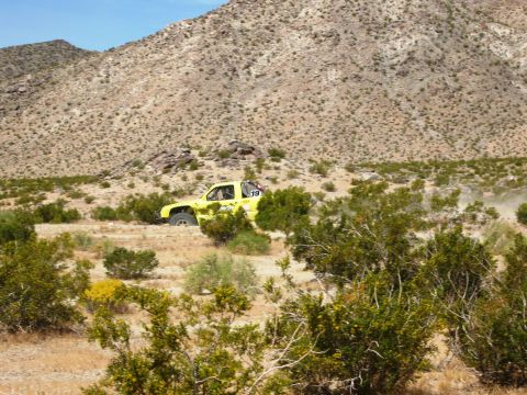 1986 Ford Bronco - A yellow off-road truck navigates a dusty desert terrain under a clear blue sky.