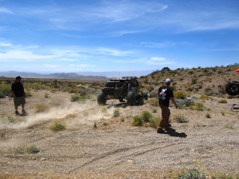 1986 Ford Bronco - Off-road buggy driving on a dusty desert track with two people watching.
