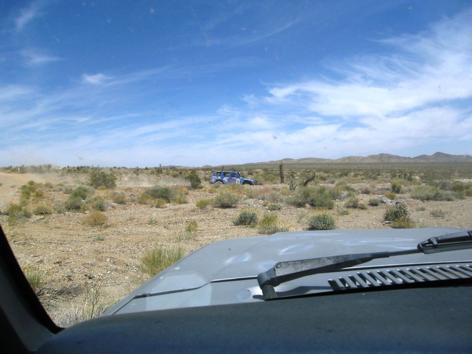 Two off-road vehicles driving on a dirt road in a desert with sparse vegetation and mountains in the background