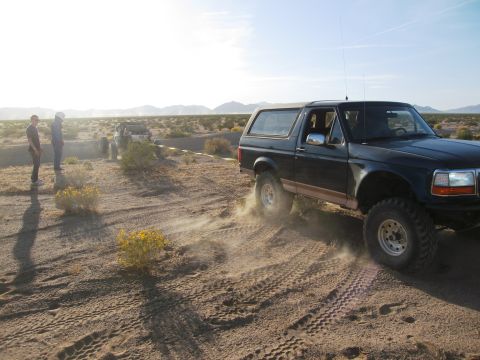 1986 Ford Bronco - A dark green Ford Bronco is seen driving off-road, kicking up dust as it goes. Two people stand in the distance watching.