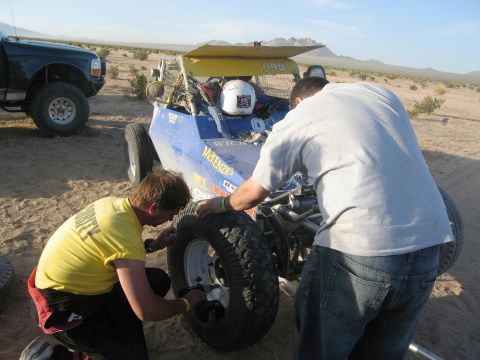 1986 Ford Bronco - Two men are working on the tire of a blue off-road buggy in a desert landscape. A pickup truck is visible in the background.