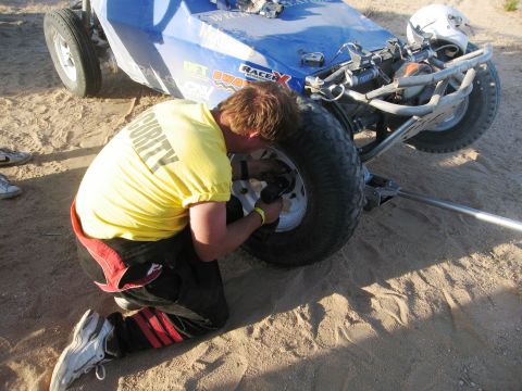 1986 Ford Bronco - A person in a yellow security shirt works on the tire of a blue off-road buggy in a sandy area.
