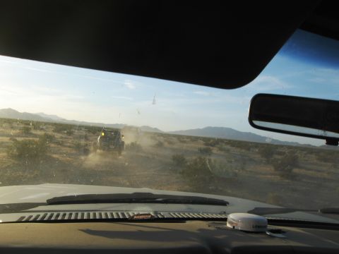 1986 Ford Bronco - Two off-road vehicles travel down a dusty desert road, seen from the perspective of another vehicle.