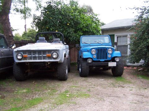 1973 Ford Bronco - Two classic off-road vehicles, a gray Ford Bronco and a blue Jeep, parked side-by-side in a yard.