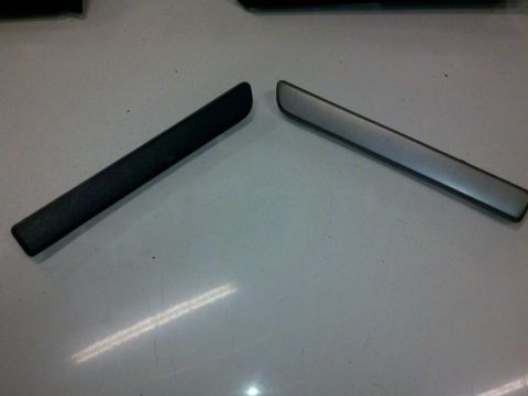 1979 Ford Bronco - Two automotive trim pieces, one black and one silver, are displayed on a flat surface.