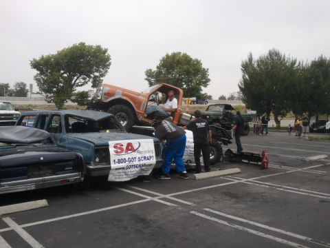 1978 Ford Bronco - An orange off-road vehicle is positioned atop a damaged blue pickup truck, which displays a recycling banner.