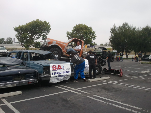 Orange off-road vehicle on crushed blue pickup truck with recycling banner