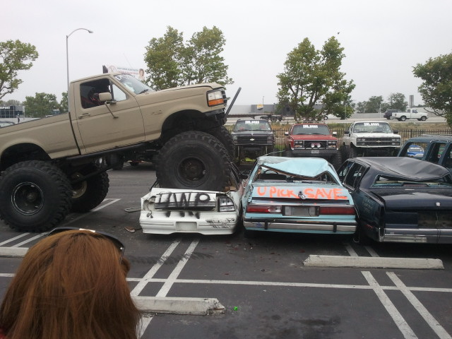 Monster truck on top of crushed cars in a parking lot. Text on cars reads 'U PICK SAVE'