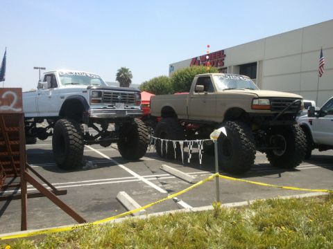 1978 Ford Bronco - Two lifted monster trucks are parked side-by-side in a lot, with a building in the background. The truck on the left appears to be from the…