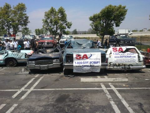 1978 Ford Bronco - A collection of damaged vehicles are gathered at a scrapyard, with one prominently displaying a recycling service banner.