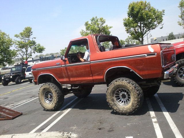 Orange 1978 Ford Bronco with lifted suspension and large tires parked in a lot. Another black Jeep is visible in the background