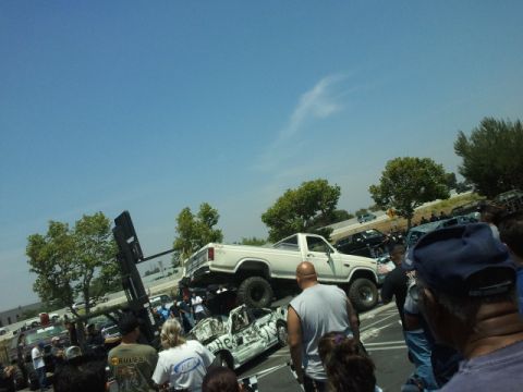 1978 Ford Bronco - A lifted white pickup truck is positioned on top of a heavily damaged car, with spectators gathered around.