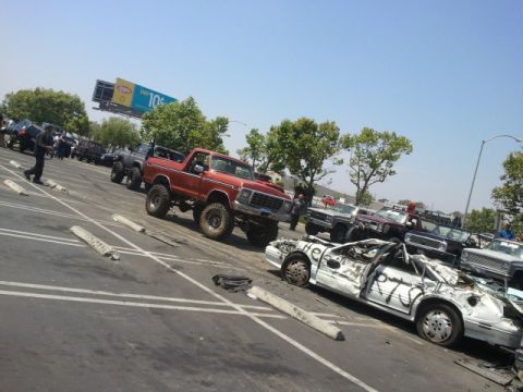 1978 Ford Bronco - A lifted red pickup truck is parked in a lot next to a severely damaged white car with graffiti on it.